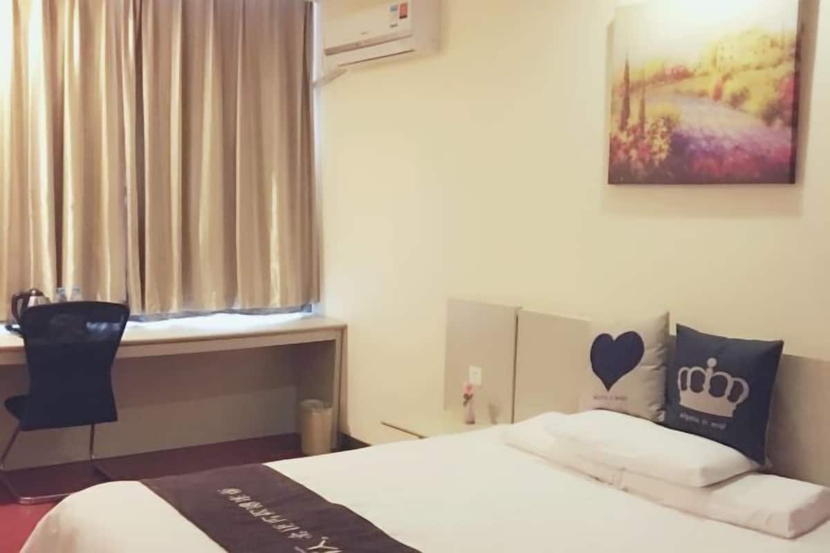 A hotel room with a bed, a desk, a chair, a window with curtains, and a painting on the wall.
