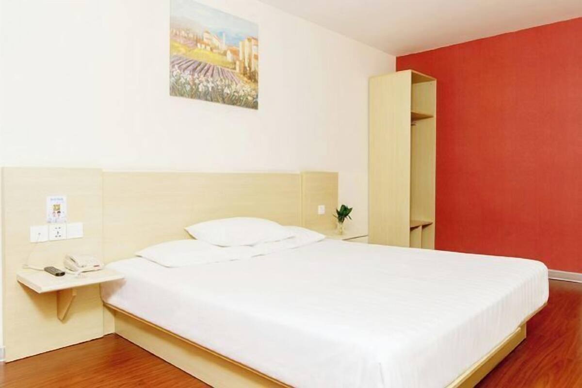 A hotel room with a bed, a bedside table with a phone, a small plant, and a painting on the wall.