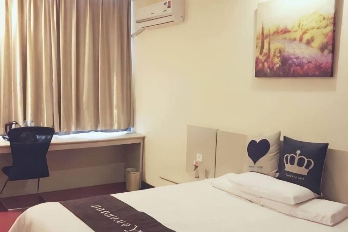 A hotel room with a bed, a desk, a chair, a window with curtains, and a painting on the wall.