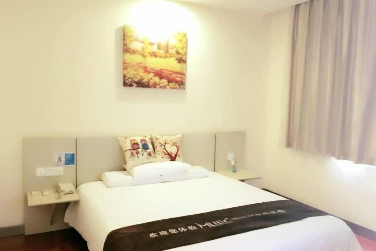 A hotel room with a bed, a bedside table with a phone, a nightstand with a lamp, and a wall-mounted artwork.