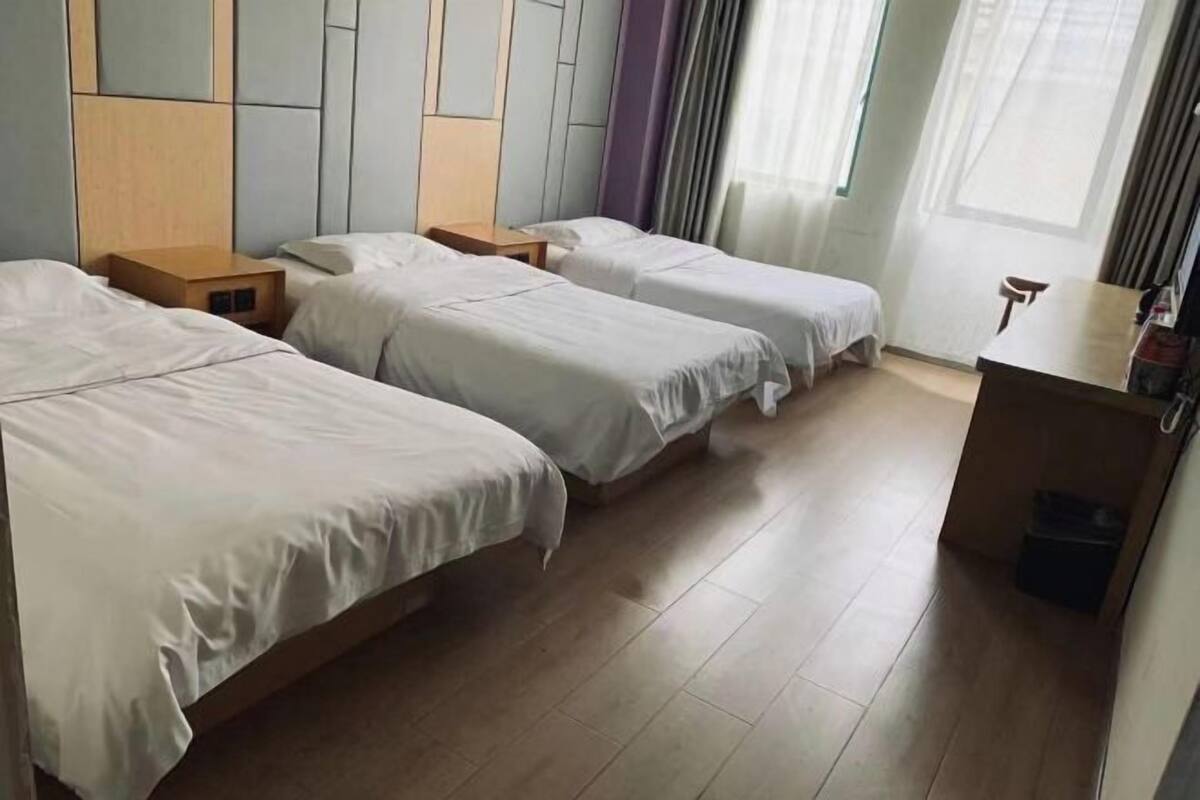 A hotel room with two single beds, a wooden headboard, a small wooden nightstand, a desk, and a window with curtains.
