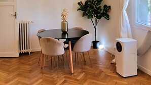 Een eetruimte met een houten visgraatvloer, een ronde tafel met vier stoelen, een moderne kroonluchter, een potplant en een radiator.