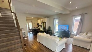 A spacious living area with a wooden floor, a white sofa, a television, and a kitchen in the background.