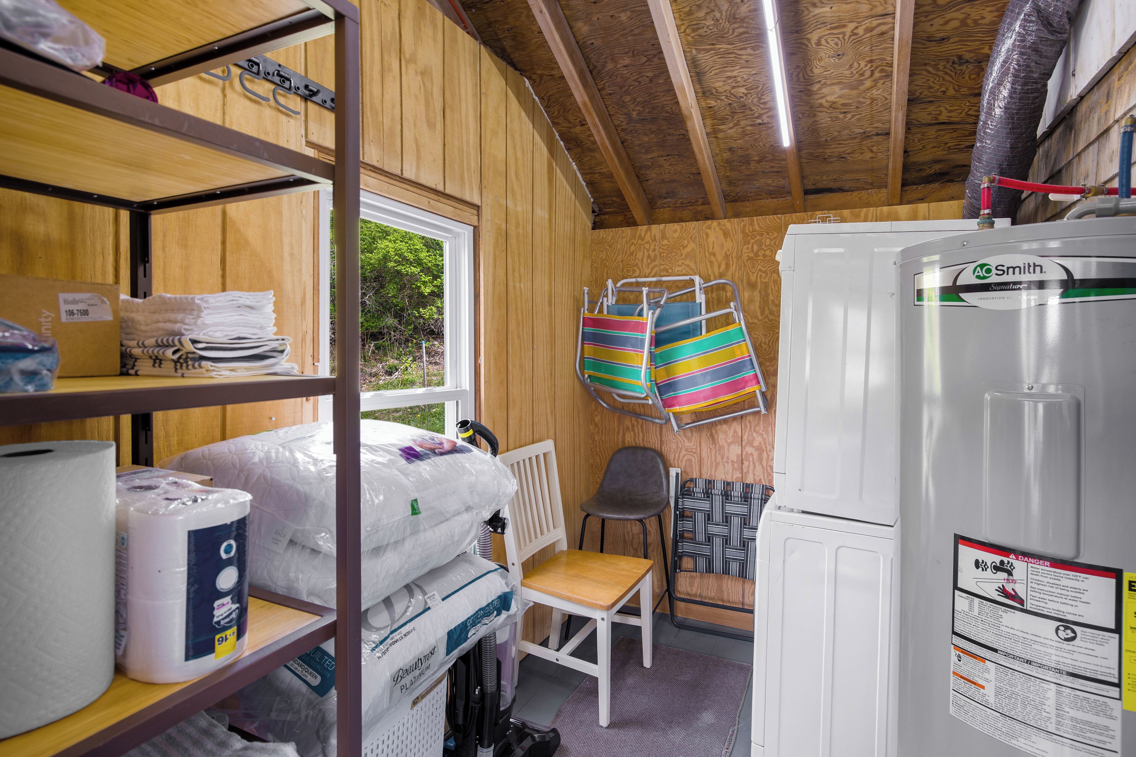 Une pièce de taille modeste aux murs et au plafond en bois, comprenant un réfrigérateur blanc, une chaise, une petite table et des étagères sur lesquelles sont disposés divers objets, notamment un grand rouleau d’essuie-tout.