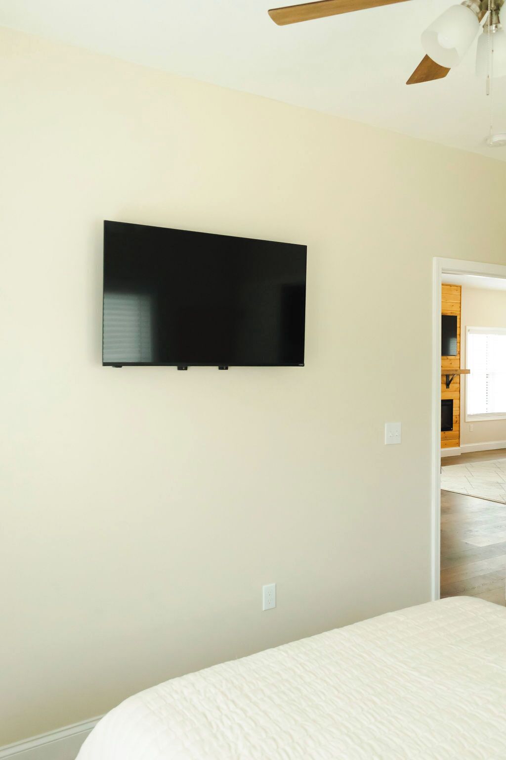 A bedroom with a flat-screen TV mounted on the wall, a bed with a white quilt, and a ceiling fan.