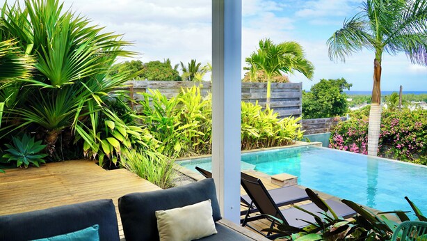 A poolside area with a wooden deck, a black sofa with cushions, and a view of the ocean. - Villa Acacias, Villa Acacias, piscine et vue panoramique (L'Étang-Salé)