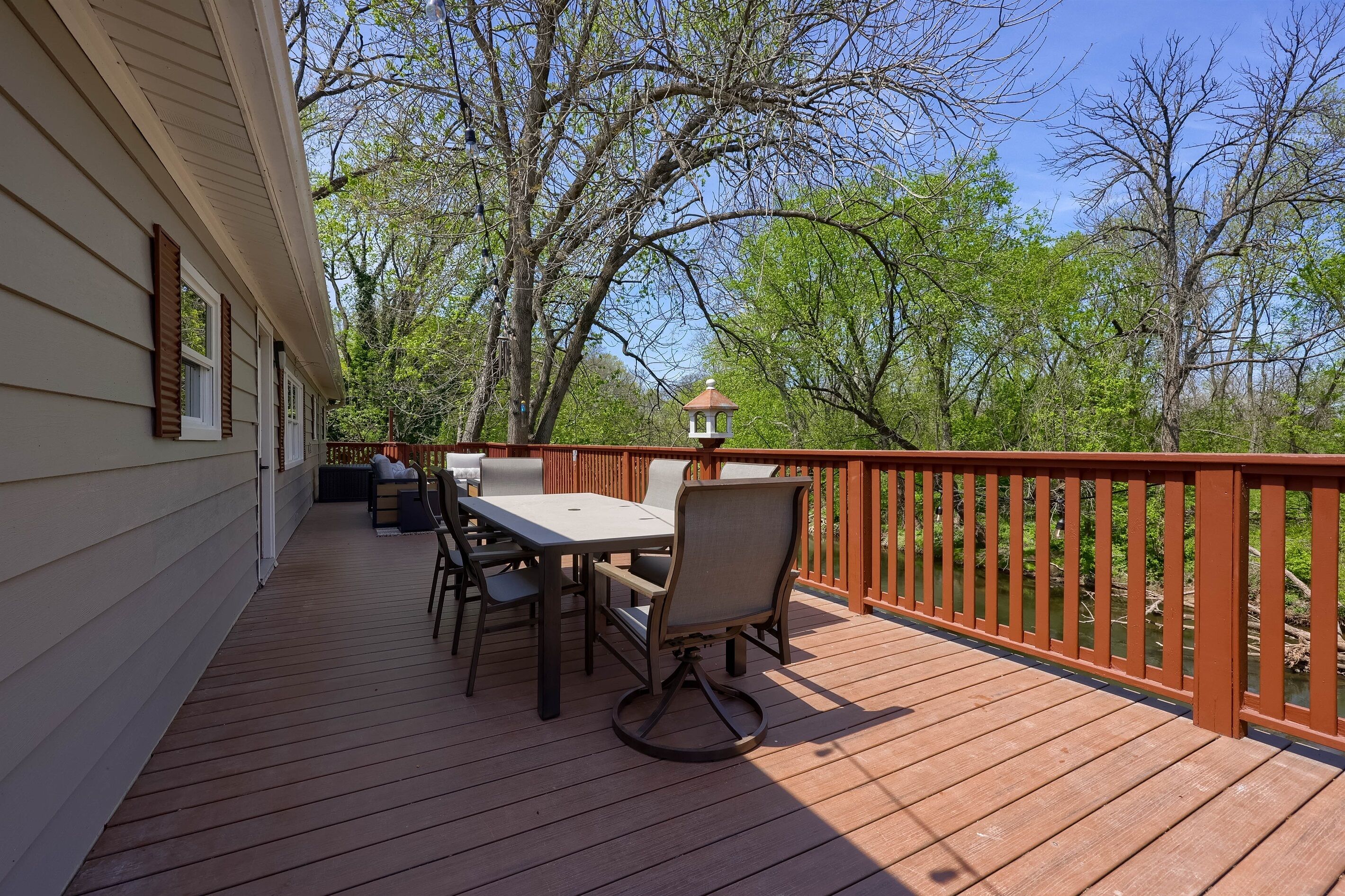 A wooden deck with a table and chairs, a lantern, and a view of trees and a fence.
