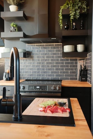 A kitchen with a modern design, featuring a black faucet, a wooden cutting board with food, and a brick backsplash.