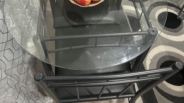 Une table de salle à manger avec un plateau en verre, sur laquelle se trouve un panier en osier rempli de bananes, d’oranges et de pommes.