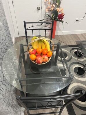A glass-top dining table with a wicker fruit basket containing bananas, oranges, and apples.