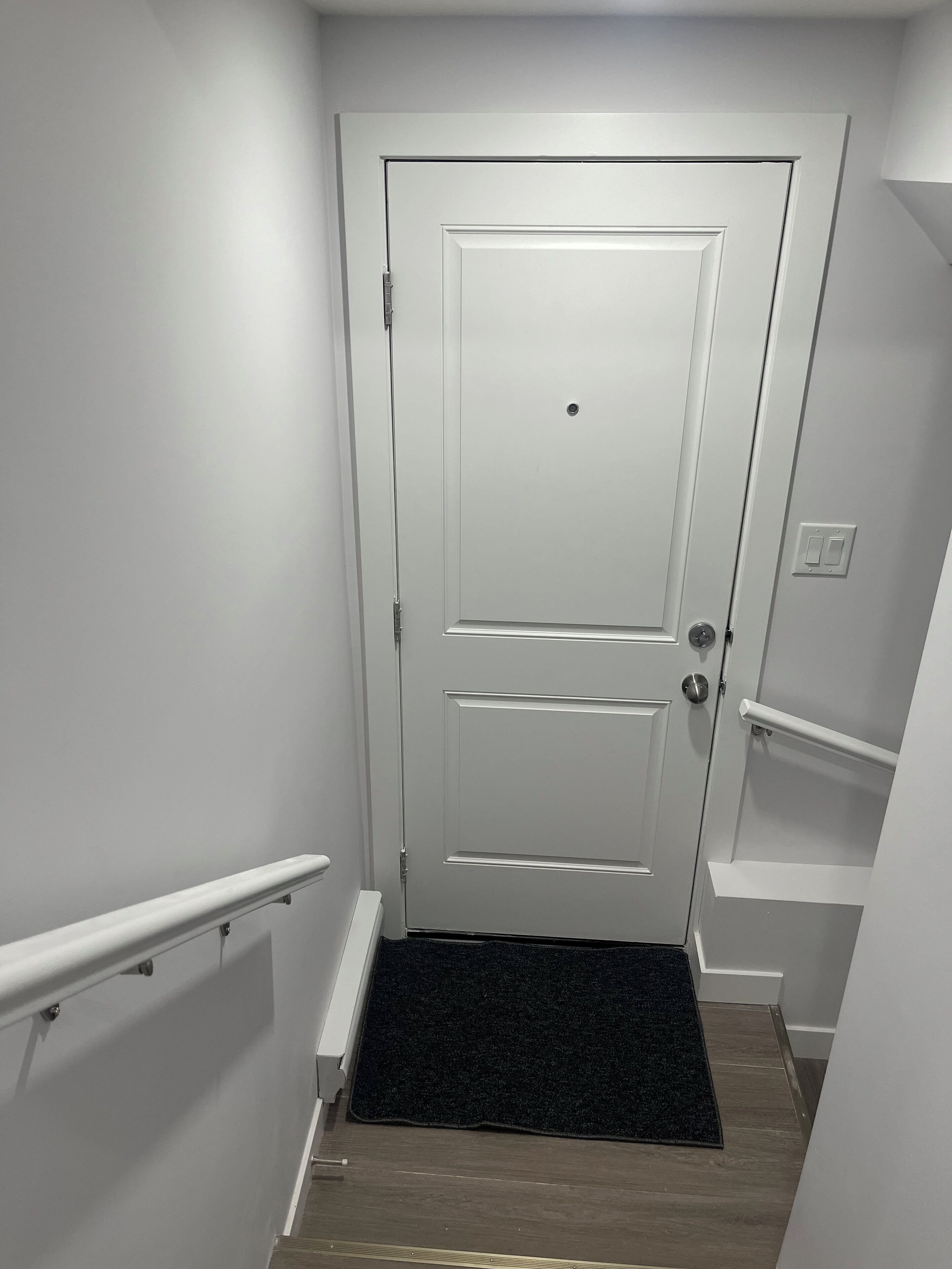 A narrow hallway with a white door, a black doormat, and a white handrail on the left.