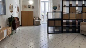A spacious living room with a high arched ceiling, a wooden wardrobe, a beige sofa, a black shelving unit, and a large window with curtains.