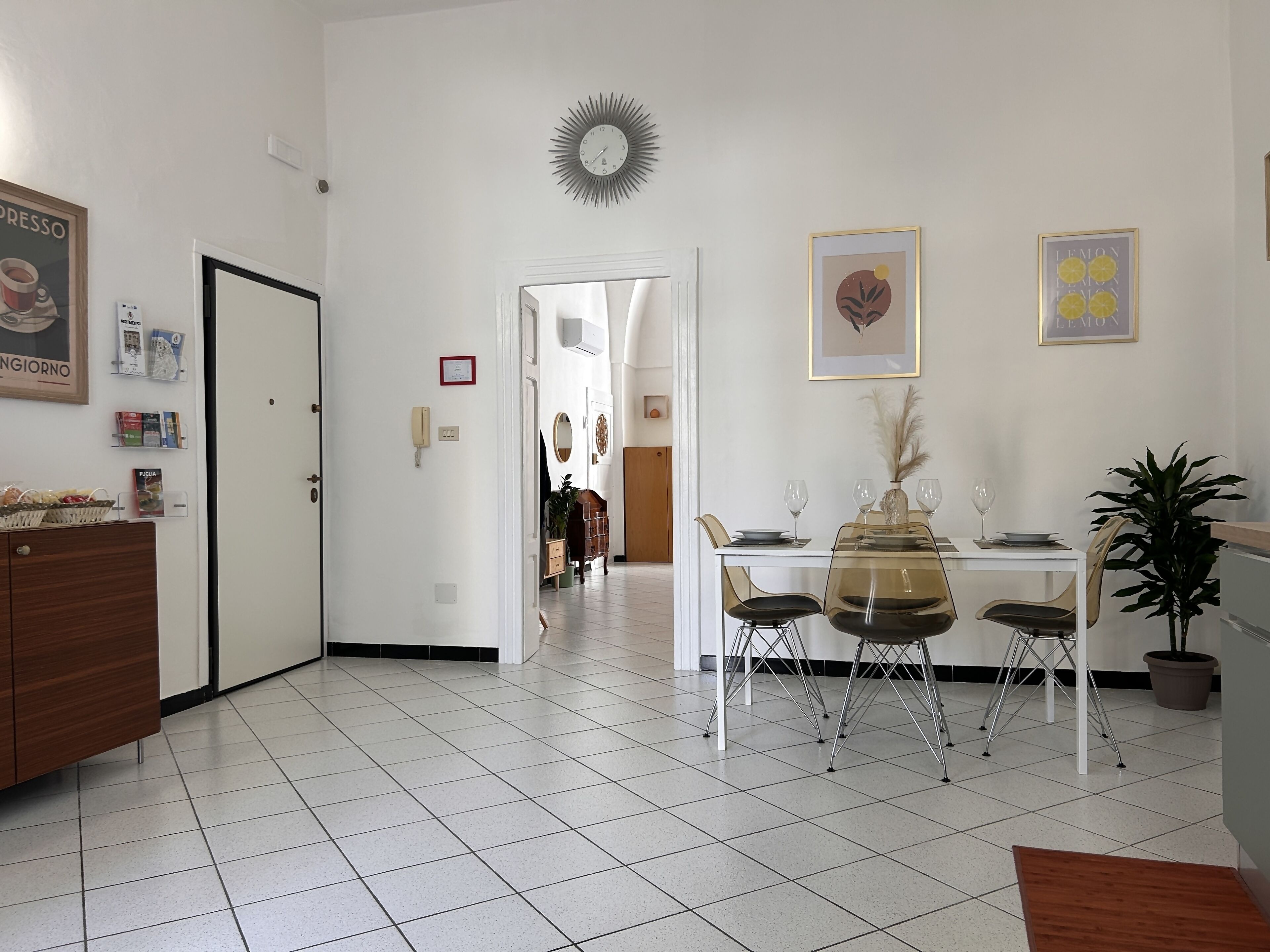 A dining area with a white table set for four, surrounded by chairs. A decorative sunburst clock hangs on the wall, and there are framed pictures above the table.