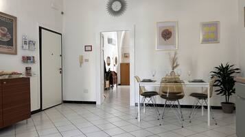 A dining area with a white table set for four, surrounded by chairs. A decorative sunburst clock hangs on the wall, and there are framed pictures above the table.