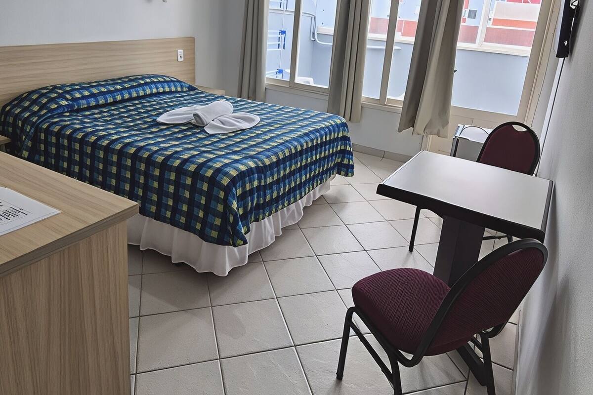 A hotel room with a bed, a desk, a chair, a window with curtains, and a ceiling light.