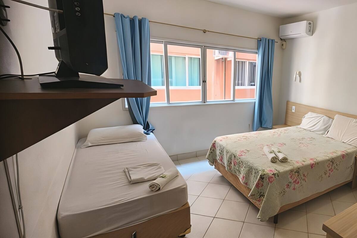 A hotel room with a bed, a desk, a computer, a window with curtains, an air conditioner, and a phone on the wall.