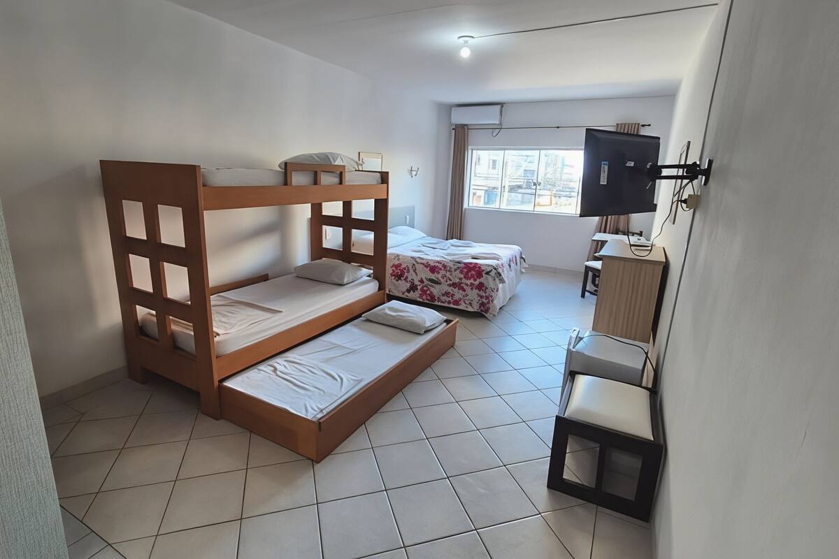 A room with a bunk bed, a single bed, a desk with a chair, and a TV mounted on the wall.