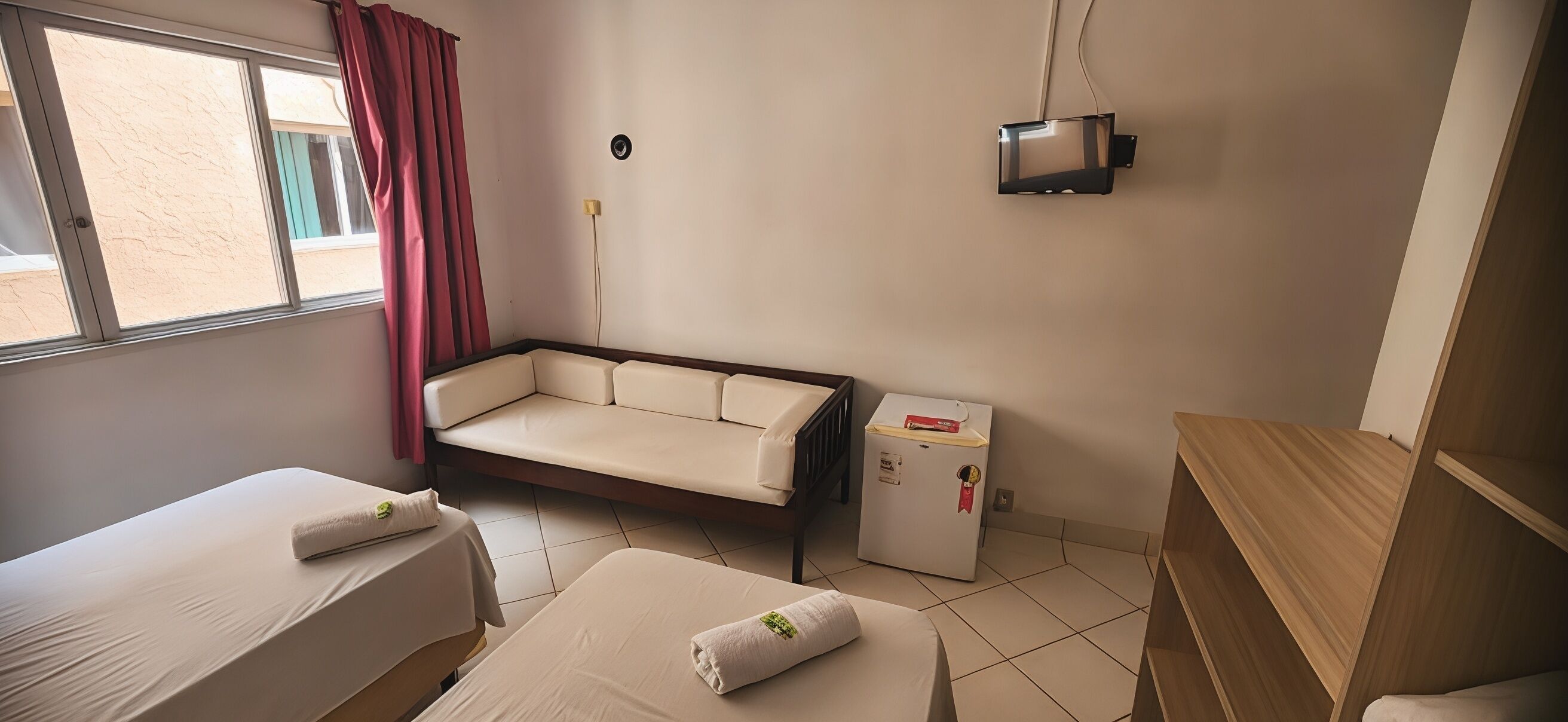 Una habitación con dos camas, un sofá, un refrigerador pequeño y una ventana con cortinas.