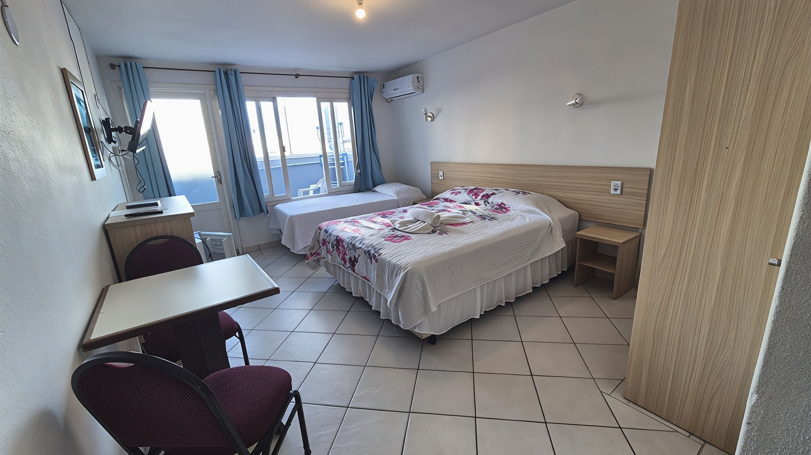 Quarto de hotel com uma cama, uma escrivaninha, uma cadeira, janela com cortinas e um ar condicionado.
