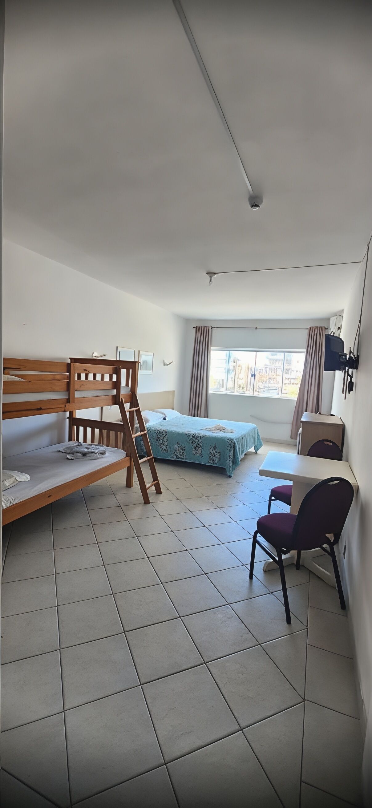 A dormitory room with bunk beds, a desk, a chair, and a window with curtains.