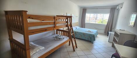 Quarto com beliches, uma escrivaninha, uma cadeira, uma cama com colcha florida e uma janela com cortinas.