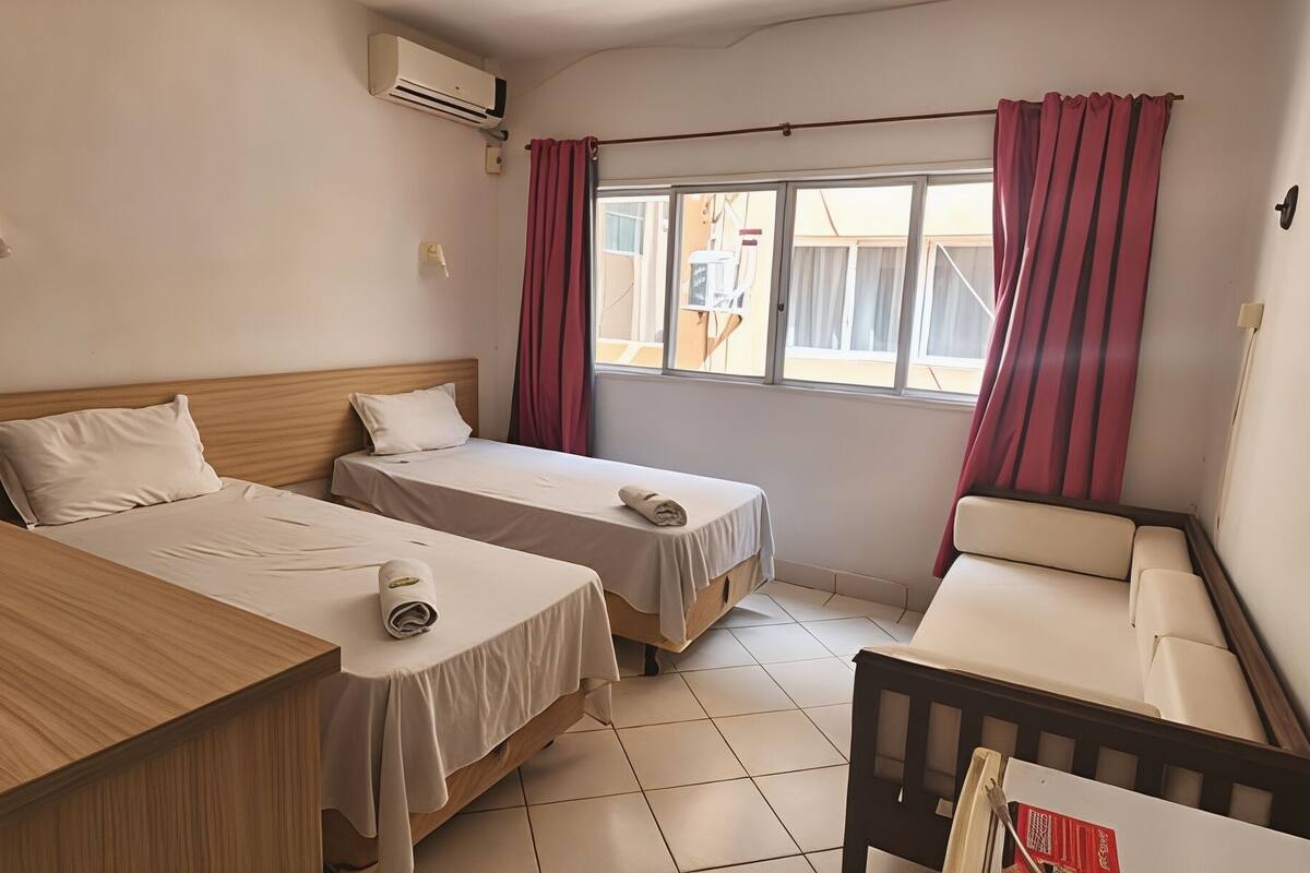 A hotel room with two beds, a wooden desk, a small sofa, and a window with curtains.
