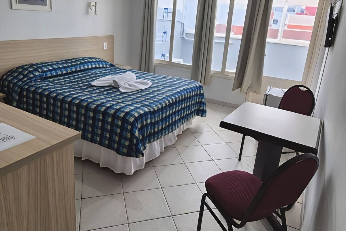 A hotel room with a bed, a desk, a chair, a window with curtains, and a ceiling light.