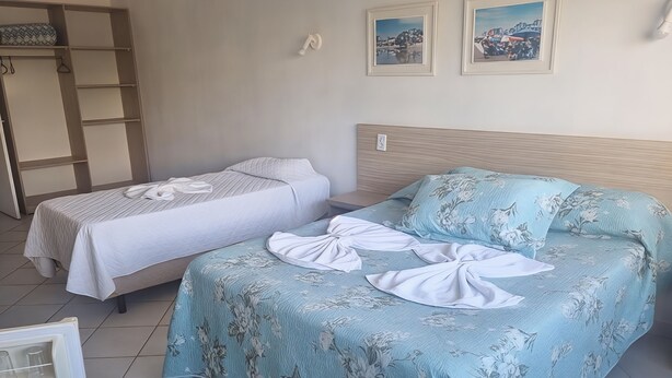 A hotel room with two beds, one with a floral bedspread and the other with a striped bedspread. There is a wardrobe with a mirror and a shelf, and two framed pictures on the wall. - Hotel Kolman (Capao da Canoa)