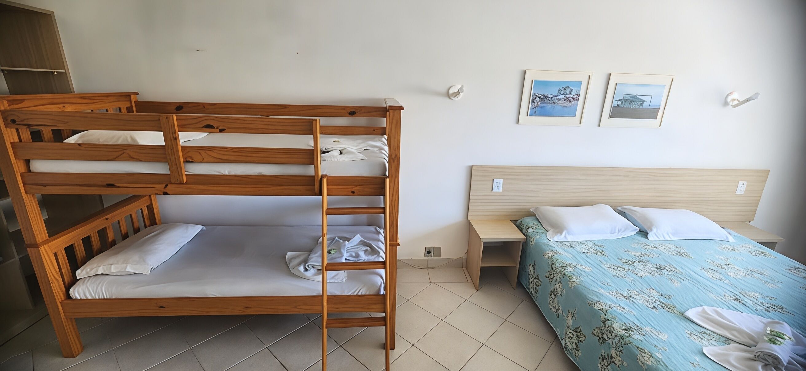 Uma cama beliche com estrutura de madeira, uma cama de solteiro com colcha florida, um criado-mudo e duas luminárias fixadas na parede.