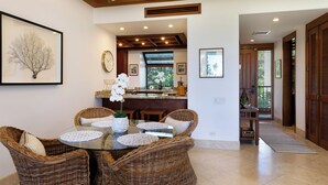 A dining area with wicker chairs and a glass table, a kitchen with a view, and a hallway leading to a door. - Mauna Lani Resort  Terrace - G301 - BEST VIEWS - Updated - Owners Club Access (Kamuela)