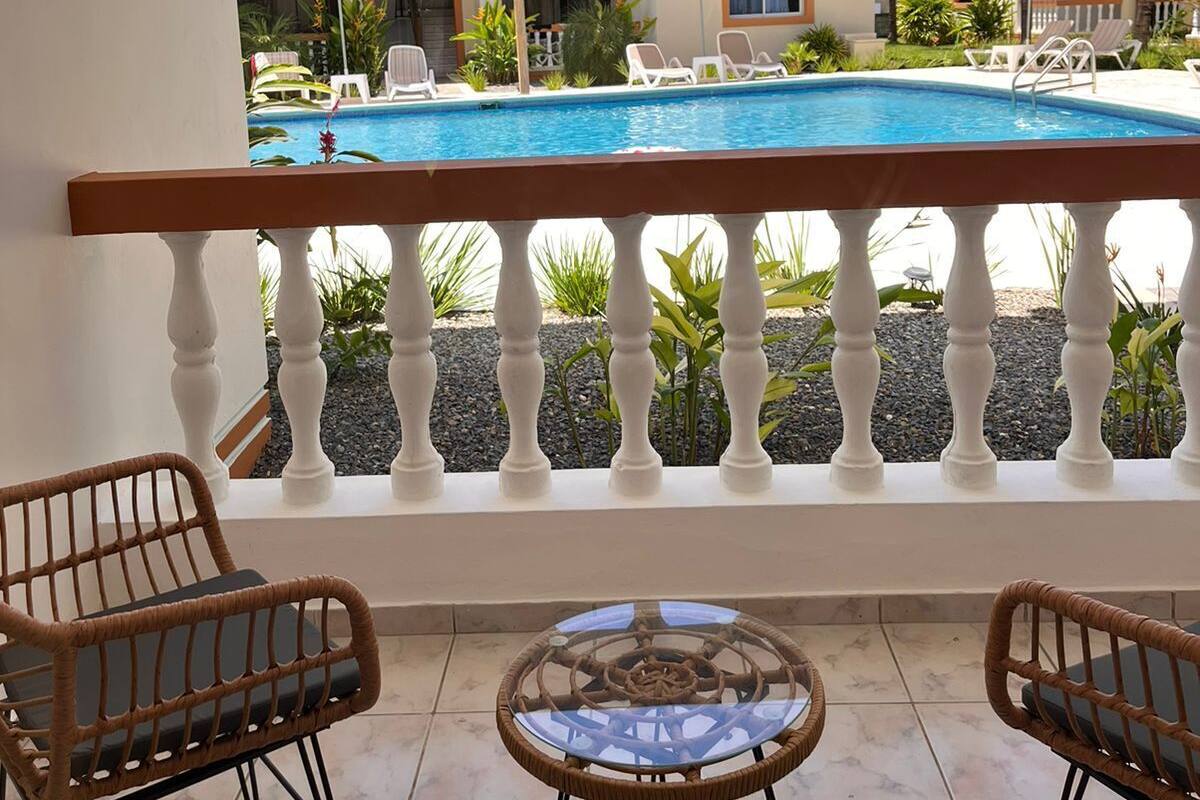 Un espace aménagé au bord de la piscine, offrant une vue sur un bâtiment dont le toit est recouvert de tuiles rouges, doté de balustrades blanches et de sièges extérieurs.