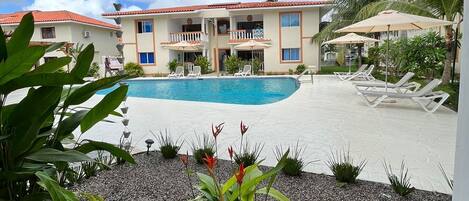 A hotel pool area with a clear view of the pool, lounge chairs, and a building with balconies.