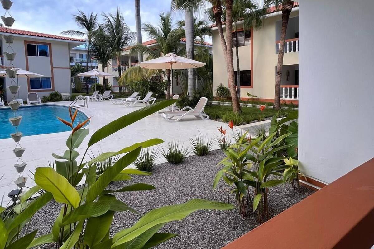 Un espace aménagé au bord de la piscine, avec des chaises longues et des parasols, entouré de palmiers et d’un bâtiment doté d’un toit en tuiles.