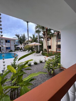 A poolside area with lounge chairs and umbrellas, surrounded by palm trees and a building with a tiled roof.