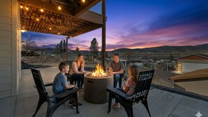 A group of people seated around a circular fire pit on an outdoor patio with a view of a residential area during twilight.