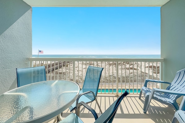 A balcony with a glass table and chairs, overlooking a beach with a fence and an American flag.