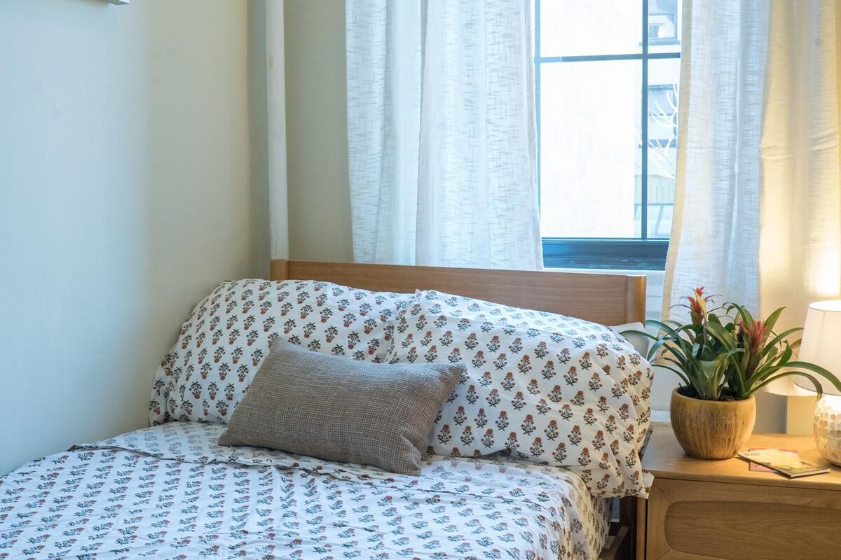 A neatly made bed with floral bedding, a bedside plant, and a lamp on a wooden nightstand.