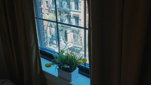 A window with a view of a cityscape, curtains partially drawn, and a potted plant with yellow flowers on the windowsill. - Interfaith 21 (New York)