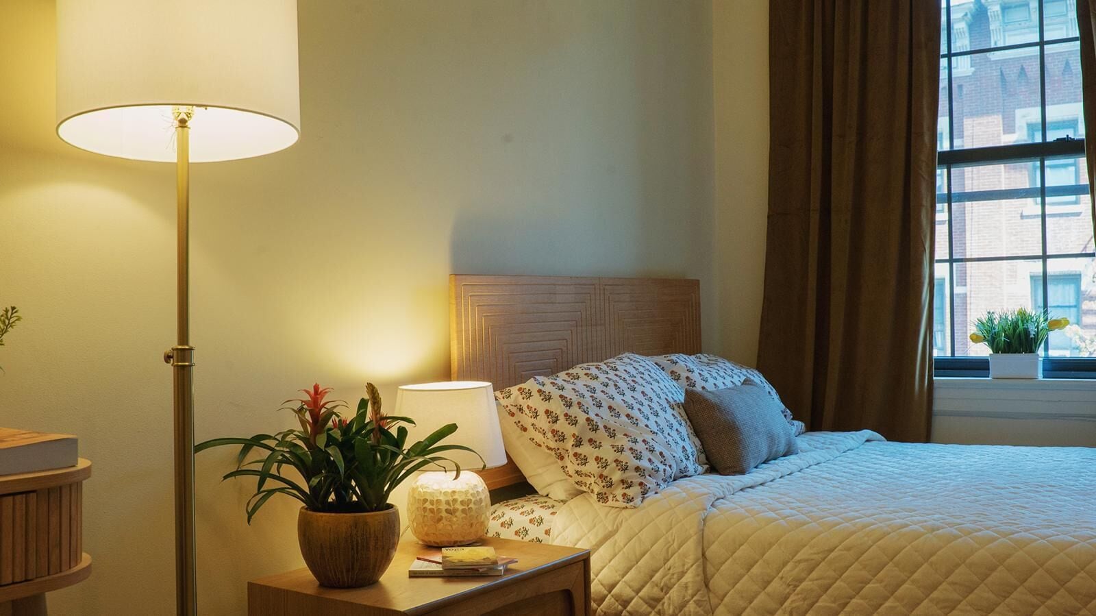 A hotel room with a bed, a bedside table with a lamp, a potted plant, and a window with curtains.