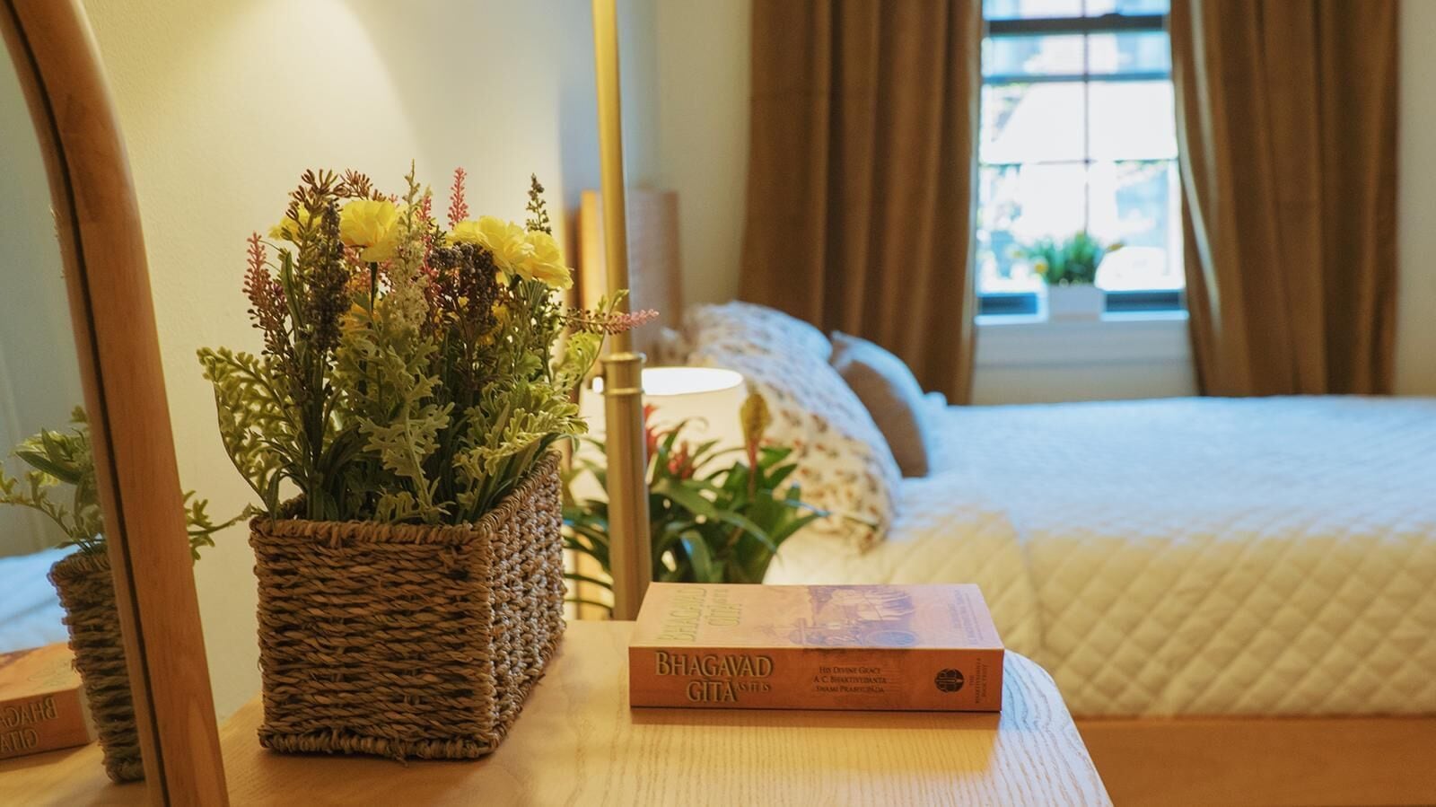 A hotel room with a bed, a bedside table with a book, a woven basket with flowers, and a window with curtains.