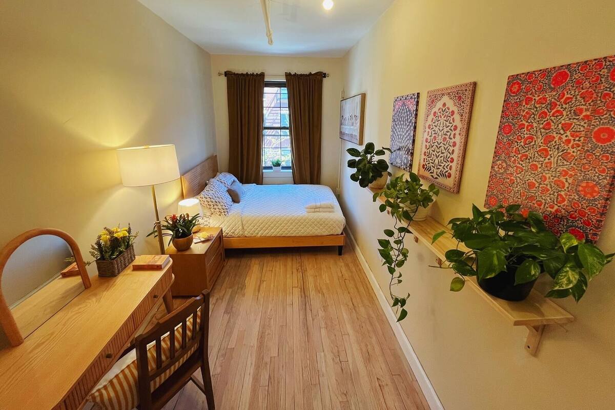 A compact hotel room with a single bed, a desk, a chair, and a small table with a lamp. There are several potted plants and decorative wall hangings.