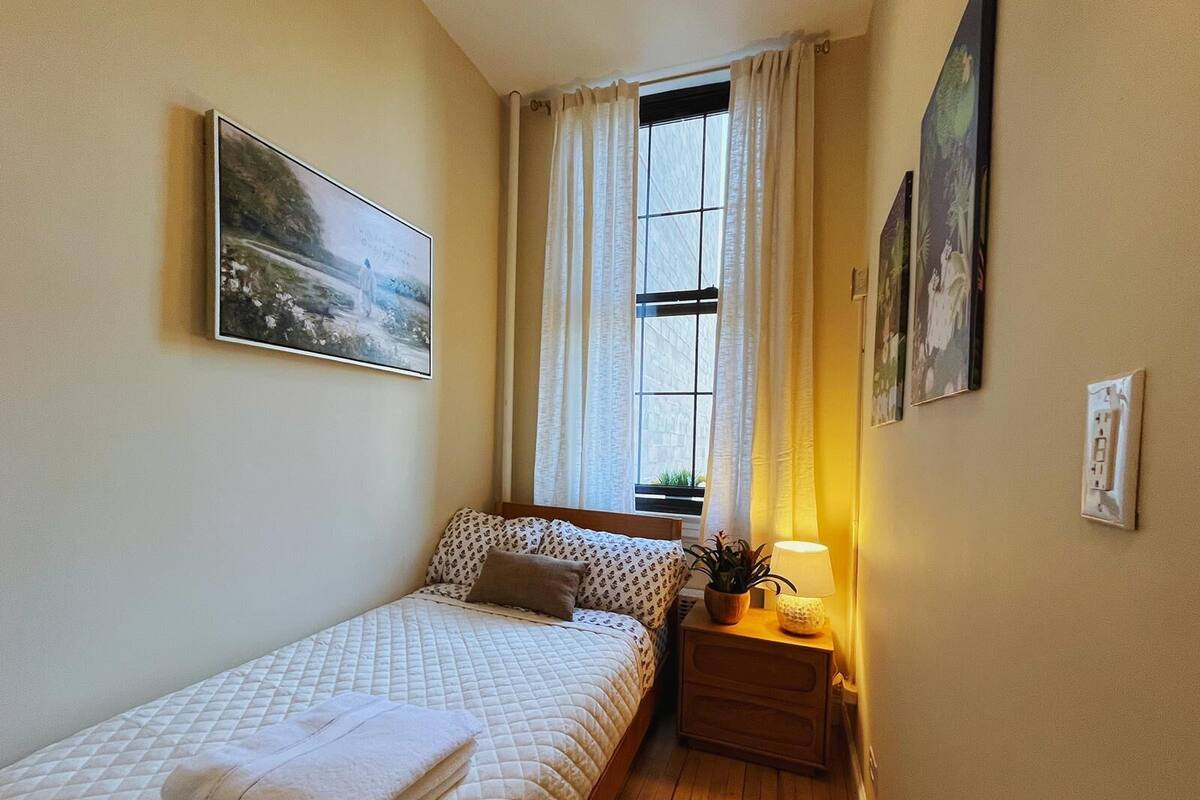 A small, cozy bedroom with a bed, a nightstand with a lamp, and a window with curtains.