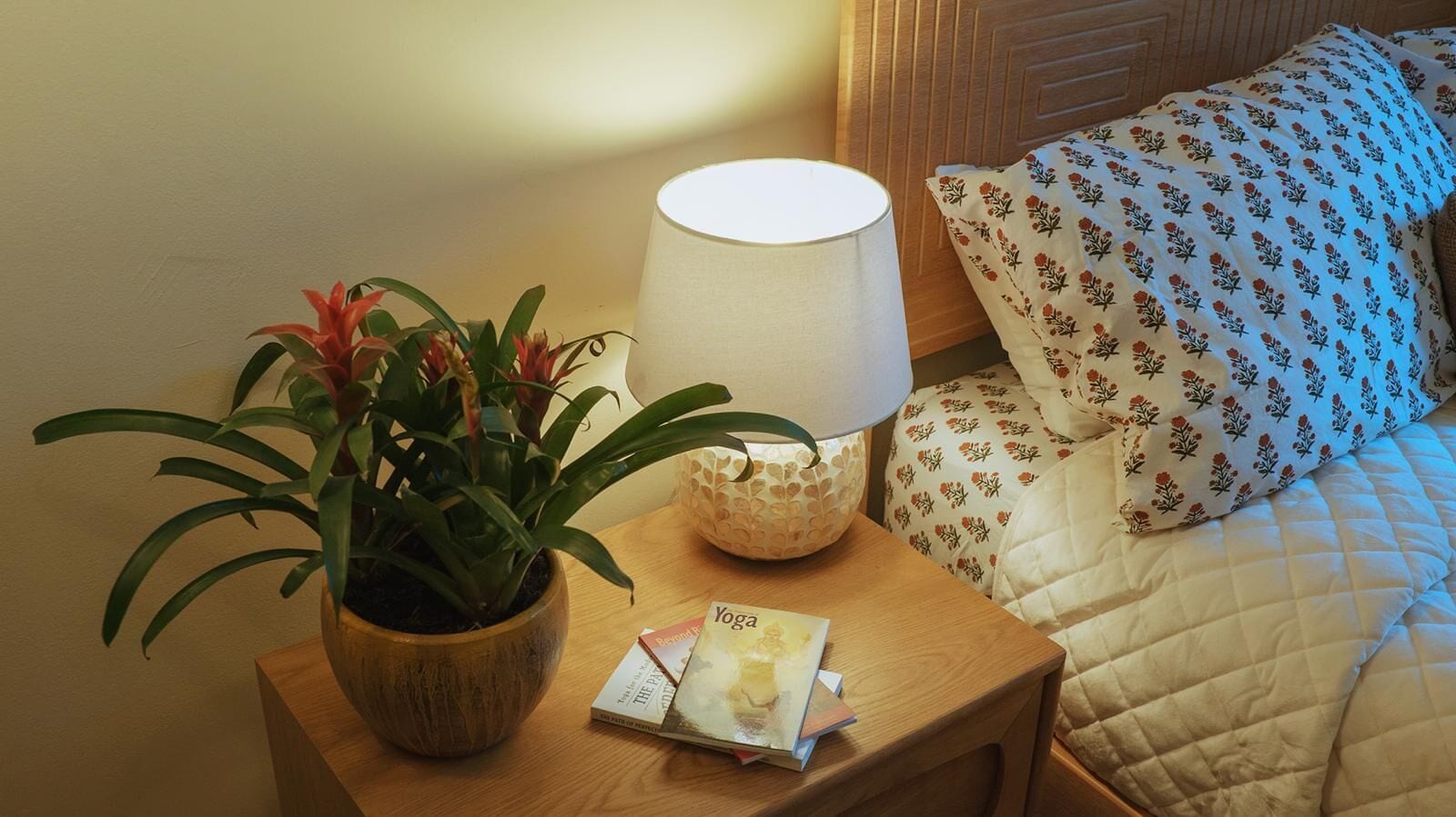 A neatly made bed with a floral patterned pillow, a bedside table with a lamp, a potted plant, and a magazine.
