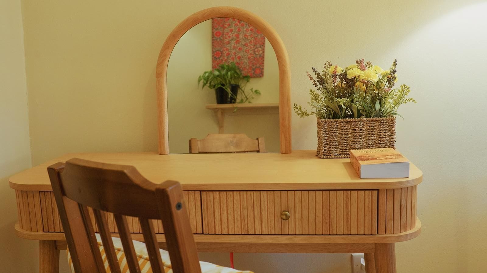 Un escritorio de madera con una silla, un espejo, un estante pequeño con una planta y una cesta llena de flores.
