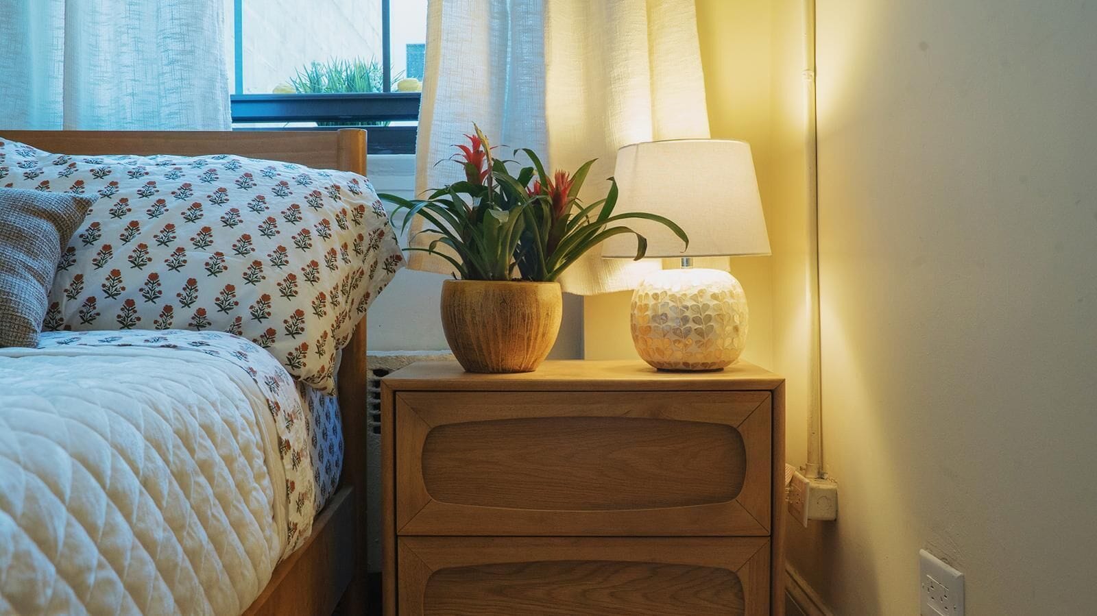 Una cama bien hecha con una colcha de estampado floral, una mesita de noche de madera con una lámpara y una planta en maceta, y una ventana con cortinas.