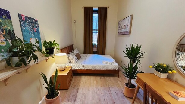 A small, well-lit bedroom with a single bed, a bedside table with a lamp, a wooden chair, and several potted plants. - Interfaith 21 (New York)