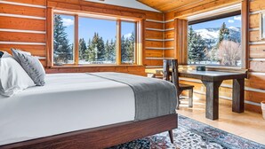 Un dormitorio tipo cabaña de madera con una cama, un escritorio y ventanas amplias que dan a una vista de la montaña.