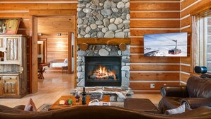 Un acogedor interior de cabaña con chimenea de piedra, paredes de madera, mobiliario de cuero y un televisor montado en la pared.