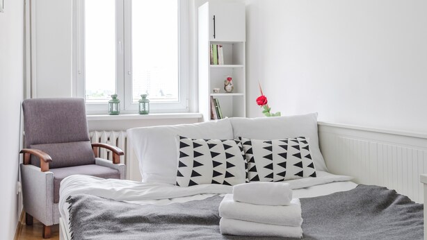 A neatly arranged bedroom with a bed, a chair, a bookshelf, and a window with blinds.