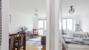 A modern, well-lit room with a wooden herringbone floor, a bed with a grey and white patterned duvet, a dining area with wooden chairs and a table, and a large window providing natural light.